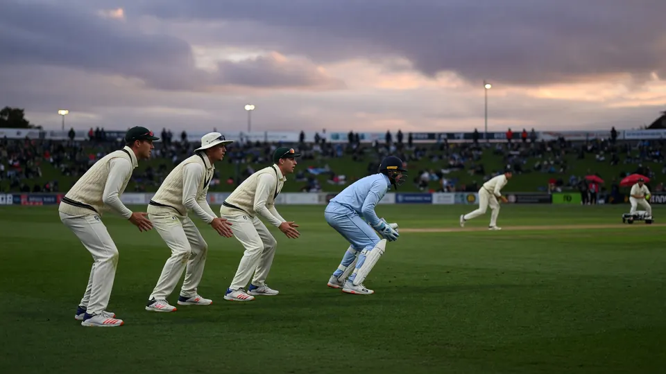 Fielders standing in a catching cordon on a green outfield under cloudy dusk light